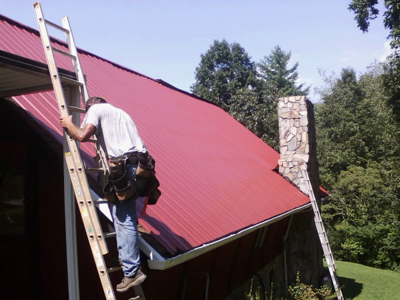 Roofer on a ladder beside a red metal roof and stone chimney — Soffit & Fascia Repair by Roofing Brothers in Ivor